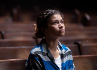 Woman with a blue striped shirt sits in a dimly lit wooden pew, gazing upward with a thoughtful expression.