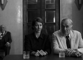 Crítica | ‘Fatherland’ discute a moderação política de Thomas Mann com direção refinada (Cannes 2026) Three adults at a dark table; woman in black, man in a light suit to her right, glasses in front; background man with ceremonial chain.