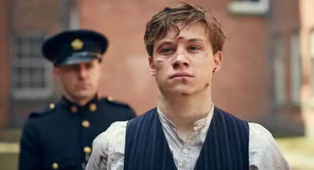 finn-cole-peaky-blinders (2) Young man with facial injuries wearing a pinstriped vest and light shirt, standing in front with a uniformed officer in the background.