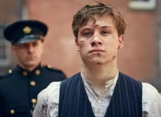 ‘Gable’: Astro de ‘Peaky Blinders’ viverá lendário medalhista olímpico em cinebiografia Young man with facial injuries wearing a pinstriped vest and light shirt, standing in front with a uniformed officer in the background.