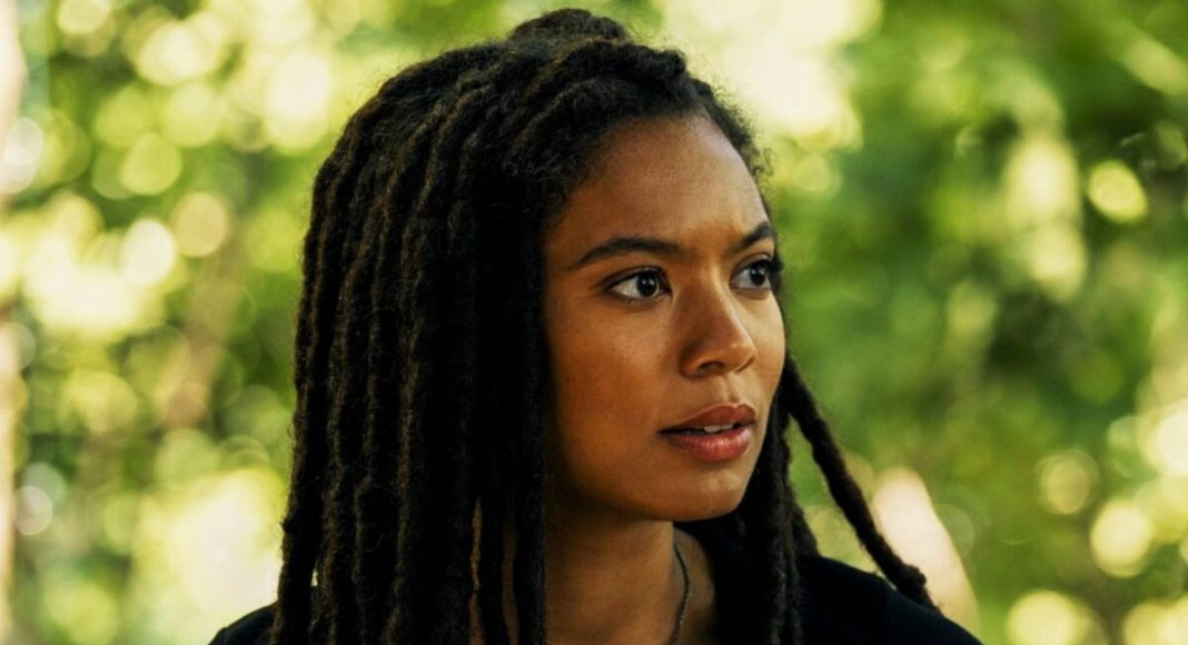 Portrait of a woman with long black dreadlocks looking to the right, outdoors with green foliage in the background.