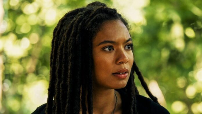 Portrait of a woman with long black dreadlocks looking to the right, outdoors with green foliage in the background.