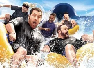 Group of friends riding a yellow inflatable raft down a water slide, splashing water around them on a sunny day.