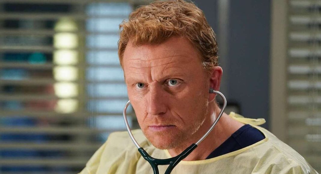 Male doctor in a light yellow protective gown with stethoscope, looking serious in a clinic setting