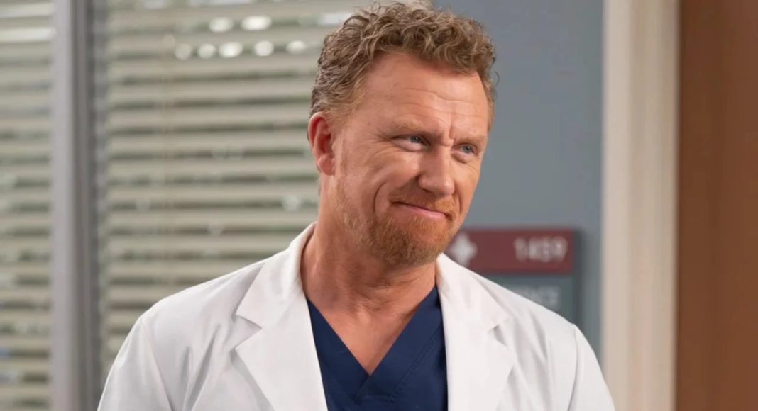 Medical professional wearing a white coat and navy scrubs, smiling in a hospital corridor.