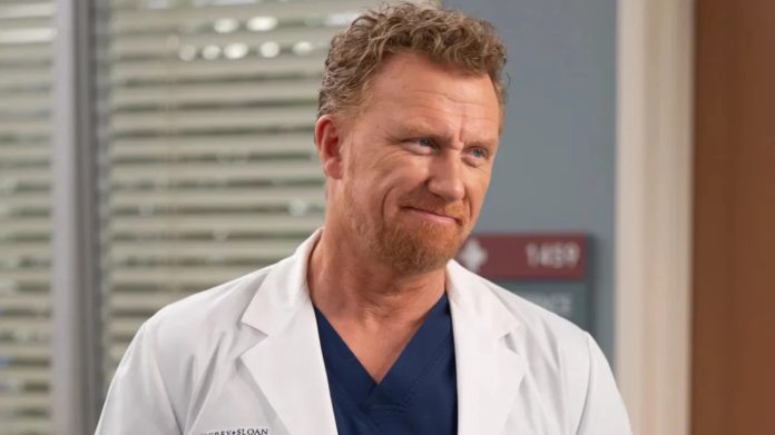 Medical professional wearing a white coat and navy scrubs, smiling in a hospital corridor.