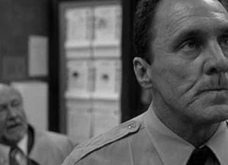 Black-and-white photo of a stern man in a shirt and tie in the foreground, with a blurred man in a suit and office shelves in the background.