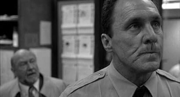 Black-and-white photo of a stern man in a shirt and tie in the foreground, with a blurred man in a suit and office shelves in the background.