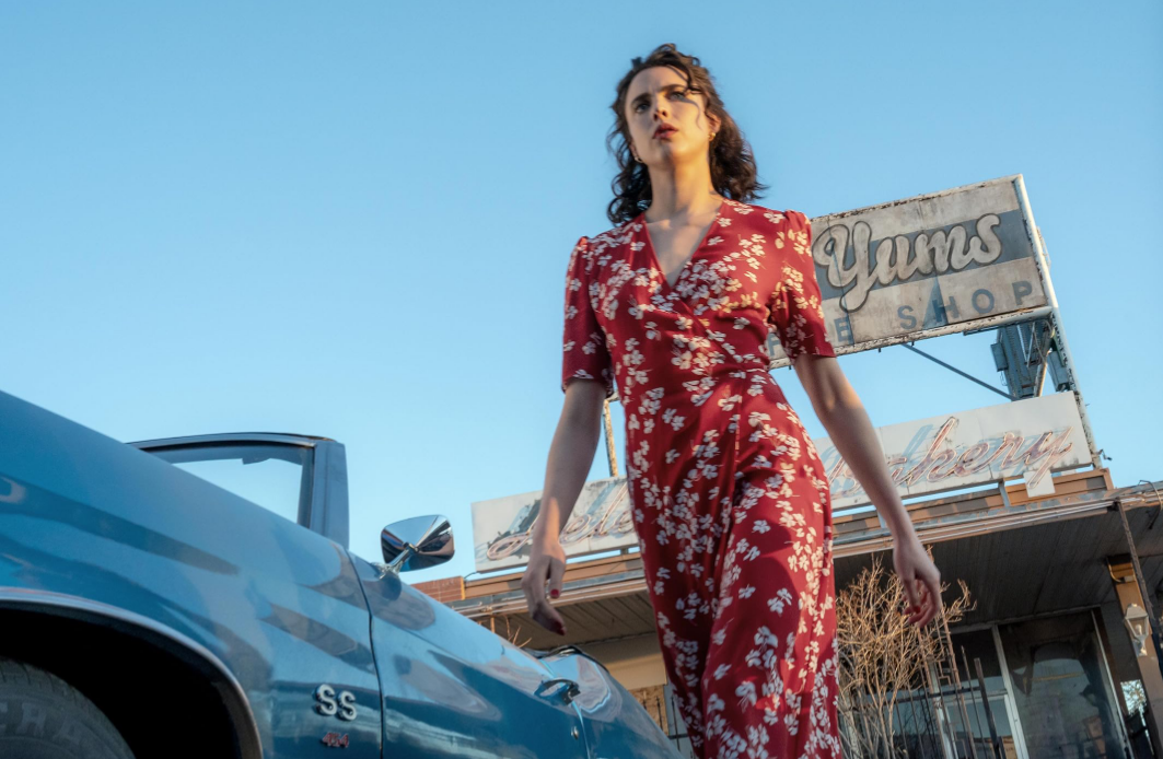 Woman in a red floral jumpsuit stands beside a blue vintage car under a clear blue sky, near an old shop sign.