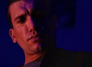 Close-up of a man in moody purple lighting, with half of his face in shadow against a dark background."