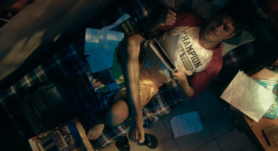 hungria1 A young man lounging on a plaid blanket bed, surrounded by papers, notebooks, and magazines in a cluttered dorm room. Overhead shot shows him reading and resting his leg on the bed.