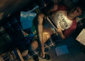 A young man lounging on a plaid blanket bed, surrounded by papers, notebooks, and magazines in a cluttered dorm room. Overhead shot shows him reading and resting his leg on the bed.