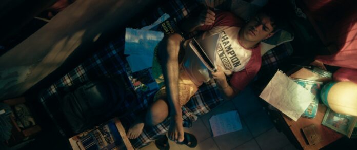 A young man lounging on a plaid blanket bed, surrounded by papers, notebooks, and magazines in a cluttered dorm room. Overhead shot shows him reading and resting his leg on the bed.