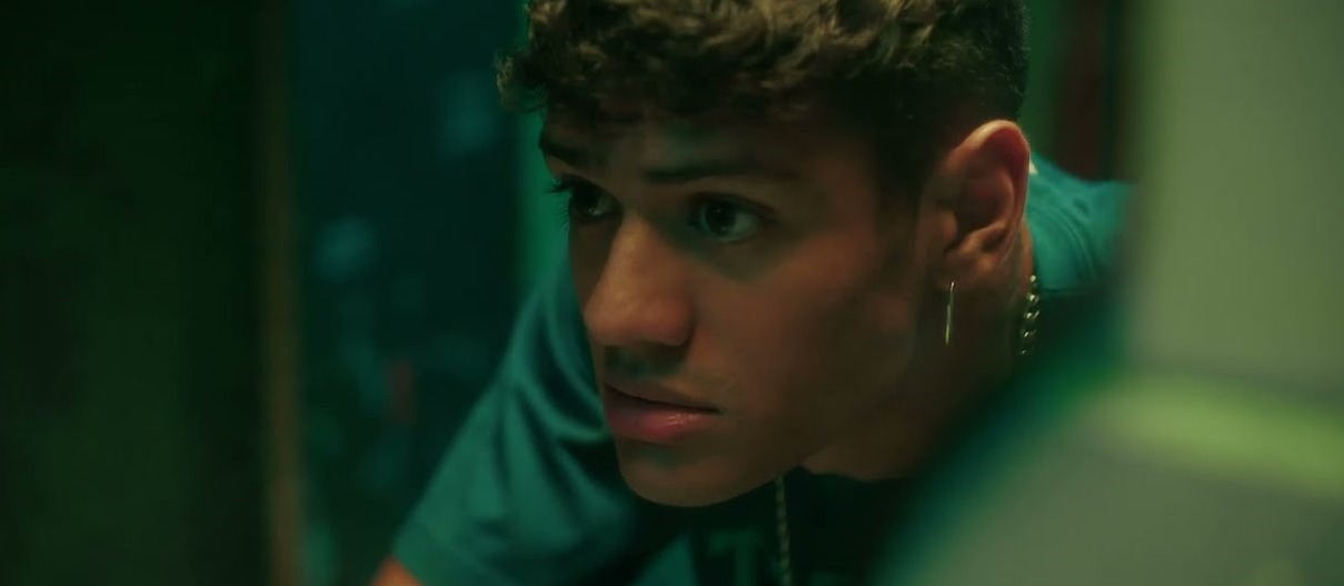 Close-up of a young man with a hoop earring and chain, gazing pensively in green-tinted lighting.