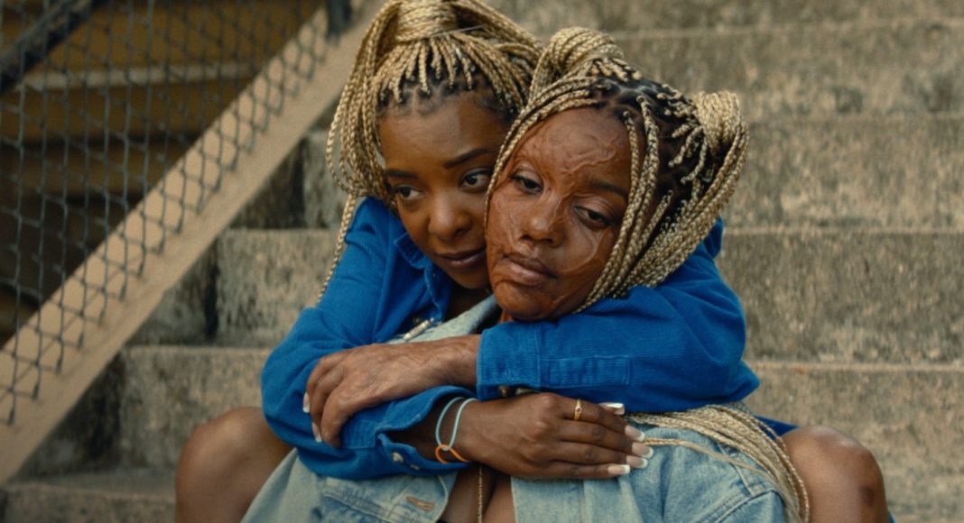 is god is 2 Two women sit on concrete stairs, embracing in a comforting hug, wearing blue jackets and denim.