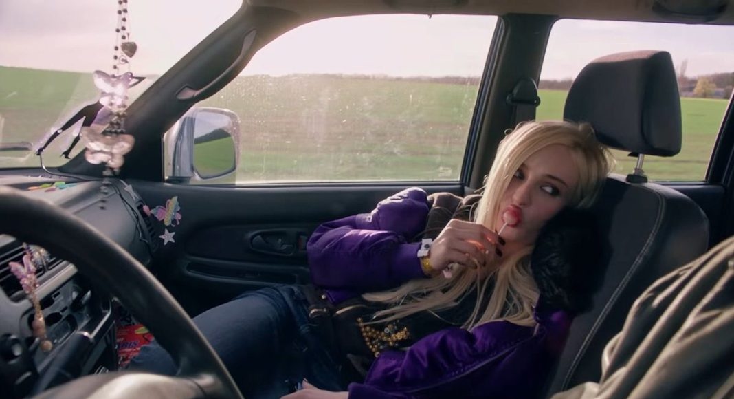 Young woman in a purple jacket lounging in a car seat, licking a pink lollipop.