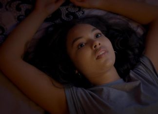 Woman lying on her back with arms raised above her head, looking upward with a thoughtful expression in dim light.