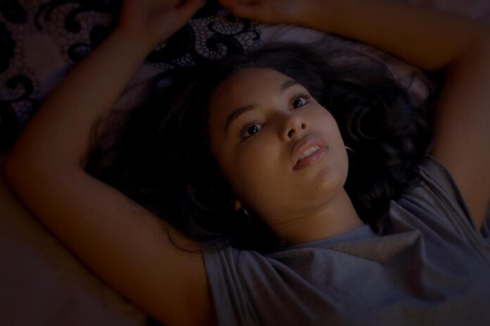 Woman lying on her back with arms raised above her head, looking upward with a thoughtful expression in dim light.