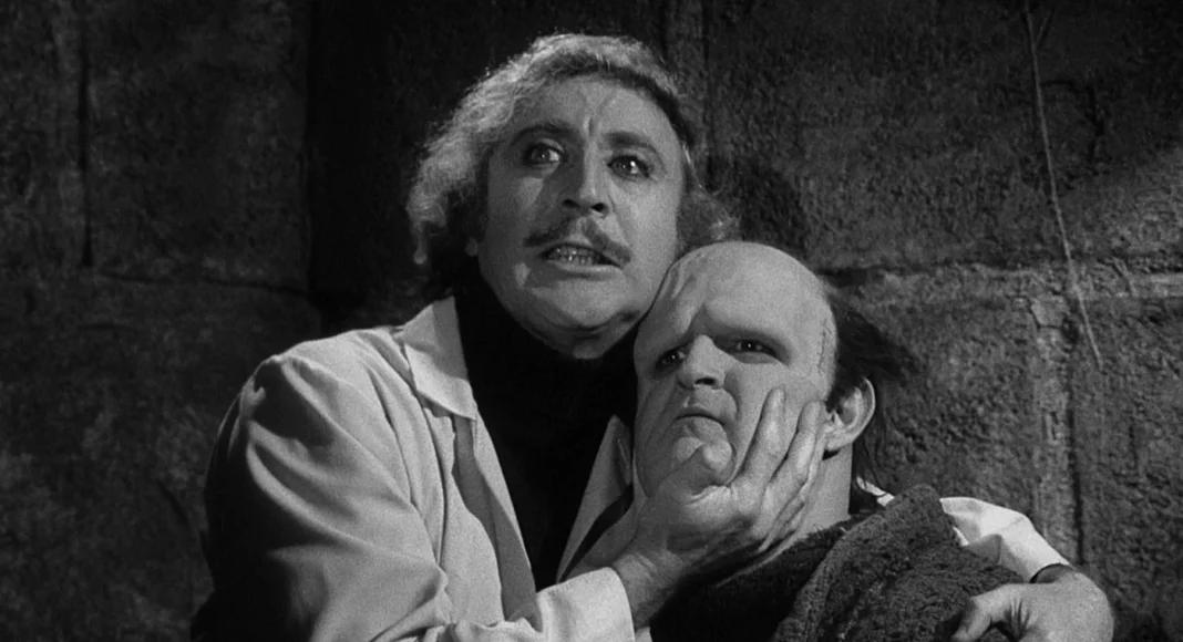 jovem frankenstein A man grips another man’s jaw in a dark, stone-walled room, creating a menacing scene.