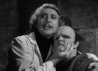 A man grips another man’s jaw in a dark, stone-walled room, creating a menacing scene.