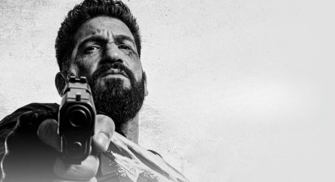 justiceiro 02 (1) Bearded man aims a handgun at the camera in a black-and-white close-up shot, low angle.