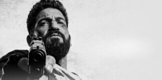 Bearded man aims a handgun at the camera in a black-and-white close-up shot, low angle.