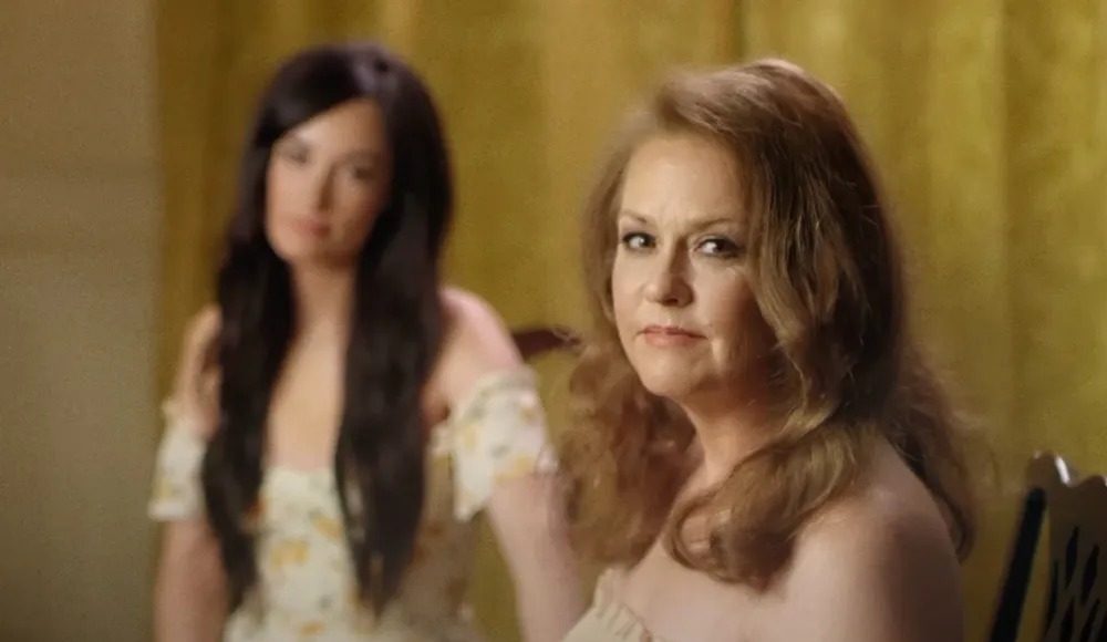 kacey musgraves 3 Two women in a softly lit room: a foreground, middle-aged woman with light brown hair wearing an off-the-shoulder cream top, looking toward the camera; a younger woman with long dark hair in a floral dress stands blurred in the background.