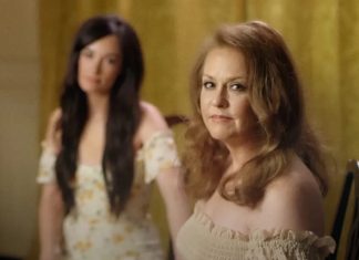 10 músicas TOCANTES para homenagear nossas mães no dia de hoje! Two women in a softly lit room: a foreground, middle-aged woman with light brown hair wearing an off-the-shoulder cream top, looking toward the camera; a younger woman with long dark hair in a floral dress stands blurred in the background.