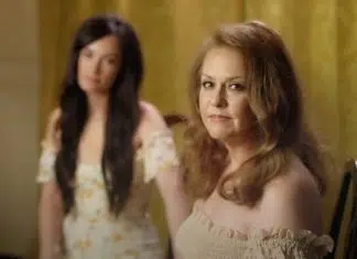10 músicas TOCANTES para homenagear nossas mães no dia de hoje! Two women in a softly lit room: a foreground, middle-aged woman with light brown hair wearing an off-the-shoulder cream top, looking toward the camera; a younger woman with long dark hair in a floral dress stands blurred in the background.