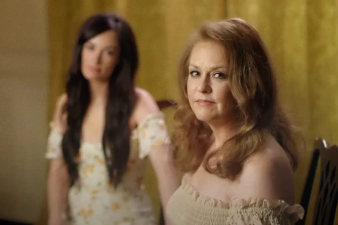 Two women in a softly lit room: a foreground, middle-aged woman with light brown hair wearing an off-the-shoulder cream top, looking toward the camera; a younger woman with long dark hair in a floral dress stands blurred in the background.