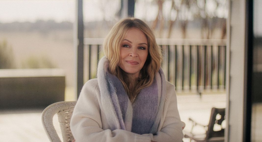 Portrait of a smiling blonde woman wearing a light sweater and lavender scarf, seated outdoors on a patio.