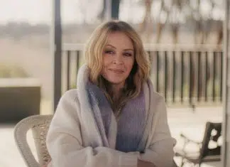Portrait of a smiling blonde woman wearing a light sweater and lavender scarf, seated outdoors on a patio.