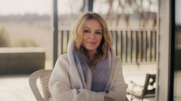 Portrait of a smiling blonde woman wearing a light sweater and lavender scarf, seated outdoors on a patio.