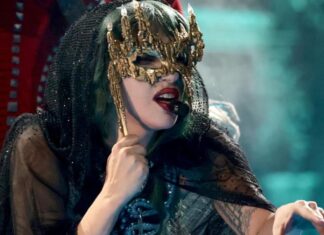 Performer wearing an ornate gold mask and black veil, singing into a headset mic on stage.