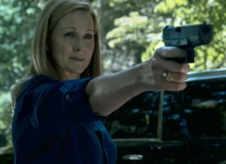 Woman in a blue shirt is pointing a handgun outward, outdoors with a dark SUV visible behind her.