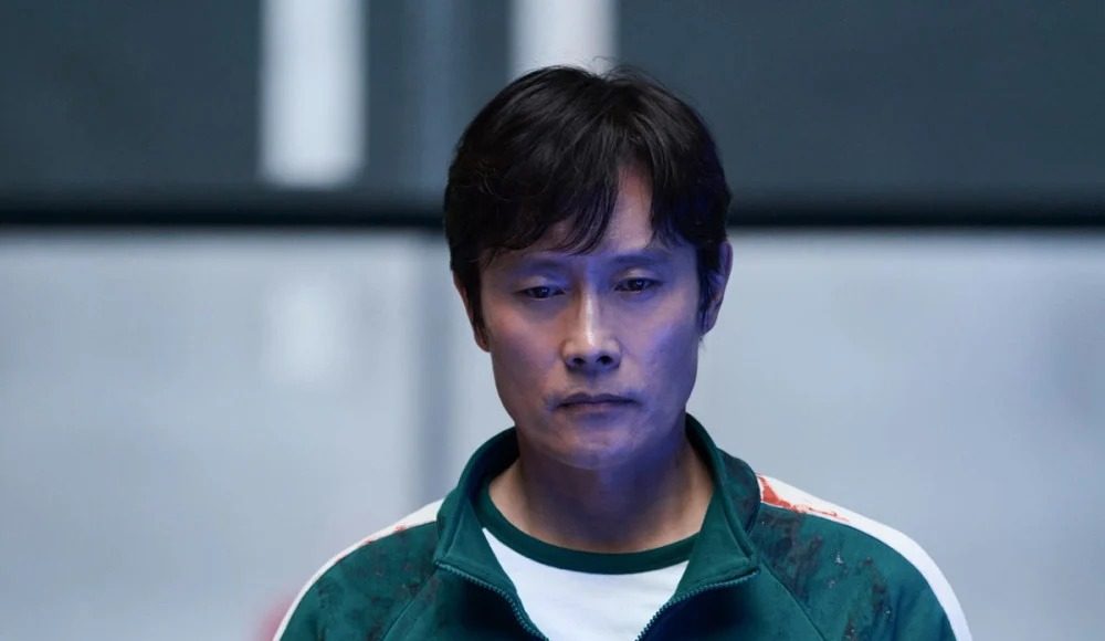 Asian man in a green zip-up jacket with white sleeves and race bibs, standing in a blue-lit indoor setting.