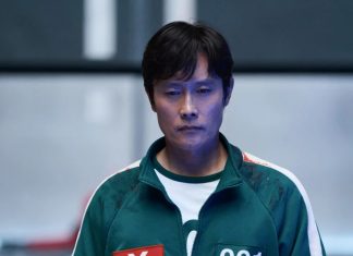 Astro de ‘Round 6’ irá estrelar o novo filme de ARTES MARCIAIS ‘Nambeol’ Asian man in a green zip-up jacket with white sleeves and race bibs, standing in a blue-lit indoor setting.