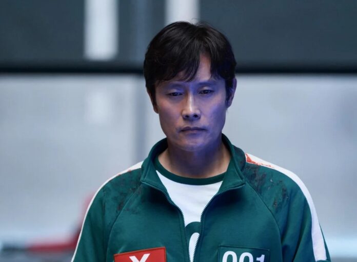 Asian man in a green zip-up jacket with white sleeves and race bibs, standing in a blue-lit indoor setting.