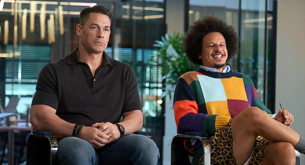Two men sit in a modern office lounge; one in a black polo, the other in a colorful patchwork sweater with leopard print shorts, both relaxed.