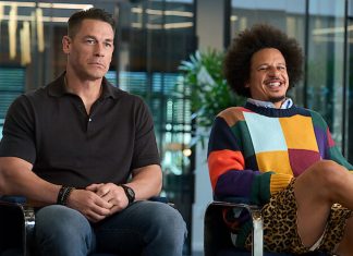 Two men sit in a modern office lounge; one in a black polo, the other in a colorful patchwork sweater with leopard print shorts, both relaxed.