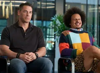 Two men sit in a modern office lounge; one in a black polo, the other in a colorful patchwork sweater with leopard print shorts, both relaxed.