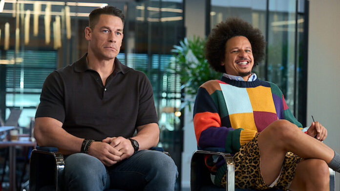 Two men sit in a modern office lounge; one in a black polo, the other in a colorful patchwork sweater with leopard print shorts, both relaxed.