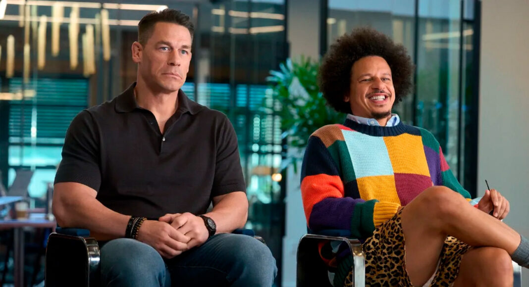Two men sit side by side in an office setting; one in a black polo looks contemplative while the other in a colorful sweater smiles and jokes.