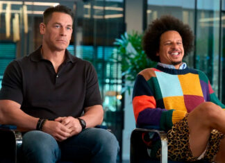 Two men sit side by side in an office setting; one in a black polo looks contemplative while the other in a colorful sweater smiles and jokes.