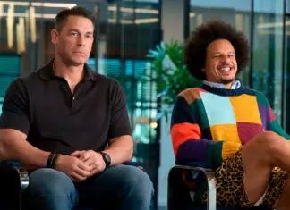 Two men sit side by side in an office setting; one in a black polo looks contemplative while the other in a colorful sweater smiles and jokes.