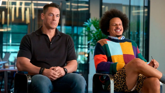 Two men sit side by side in an office setting; one in a black polo looks contemplative while the other in a colorful sweater smiles and jokes.