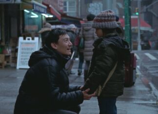 Man in a black puffer jacket kneels on a rainy city street, holding hands with a standing child wearing a knit hat.