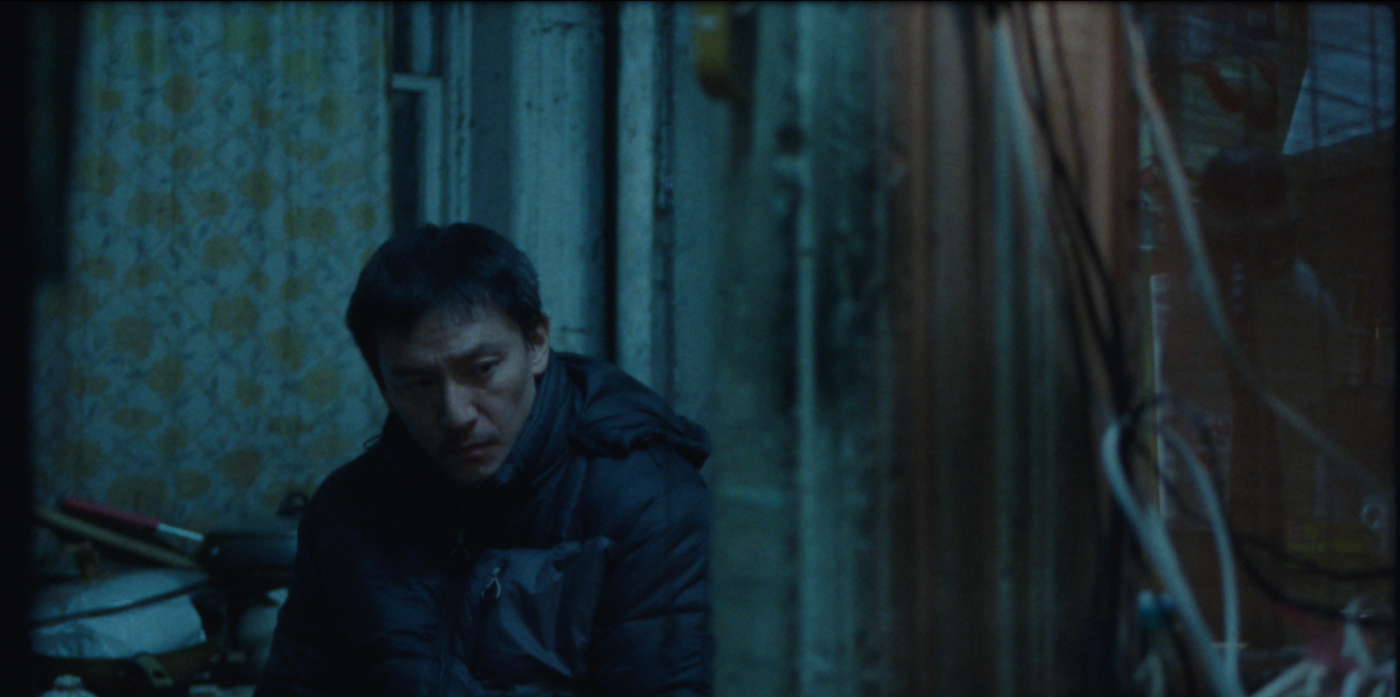 Man in a dark jacket sits in a dim, blue-lit alley, looking down with a weary expression.