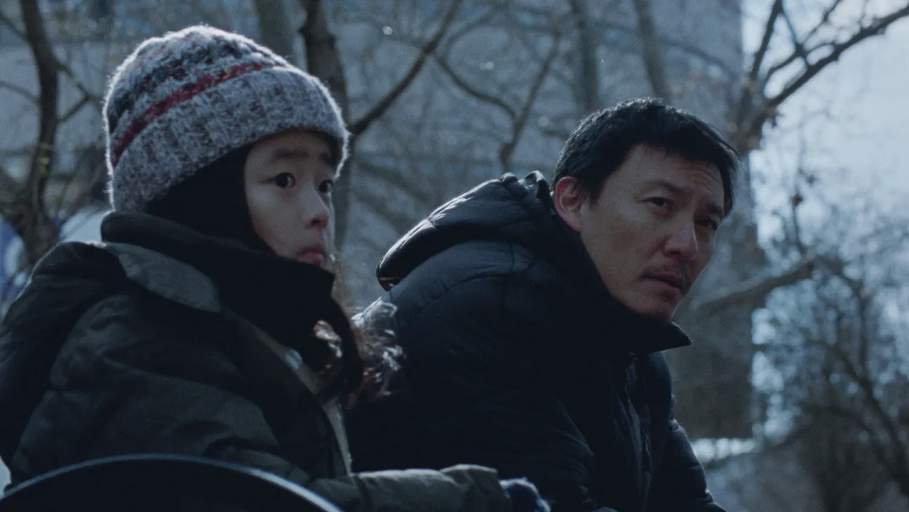 Two people in heavy winter coats sit outside, with bare trees in the background, looking concerned.