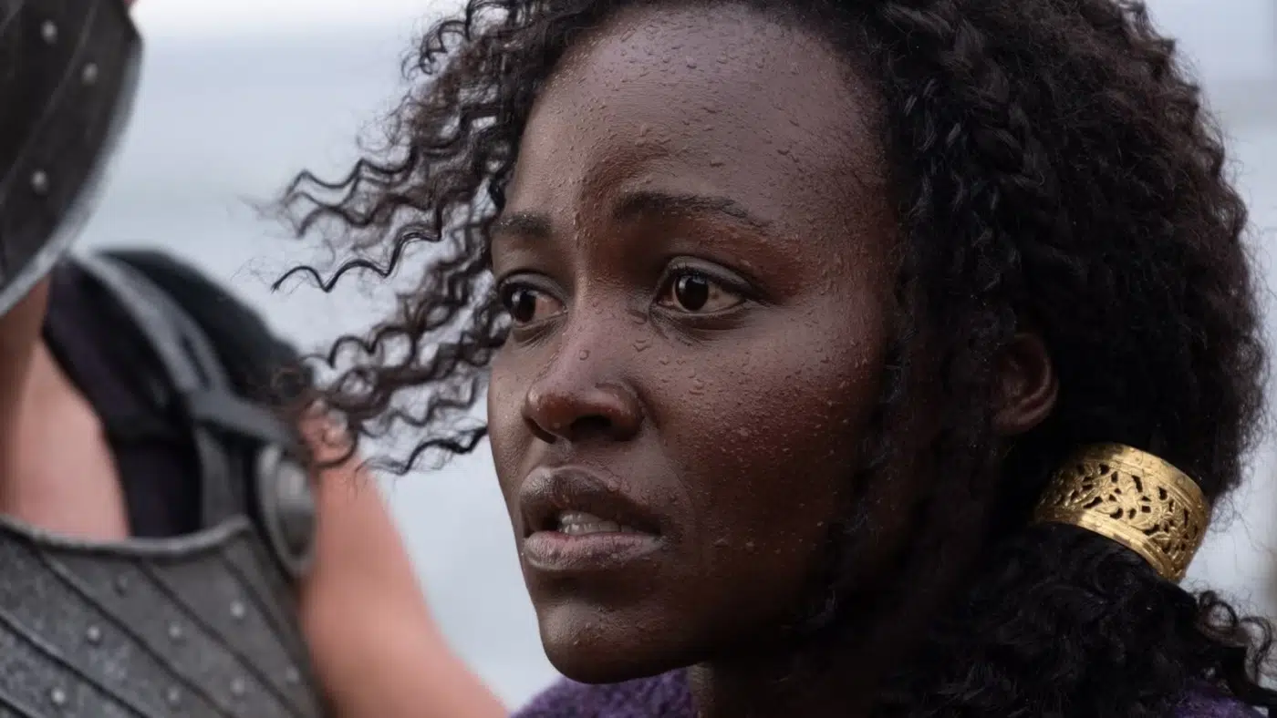 Close-up of a dark-skinned woman with wet skin and curly hair, looking distressed at a figure in armor to her left, wearing a gold hair cuff.